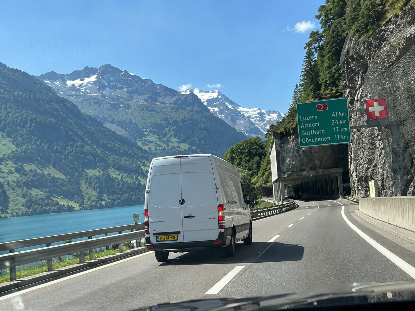 Nederlandse bestelbus met geel kenteken op een Zwitserse bergweg langs een bergmeer en besneeuwde Alpen.